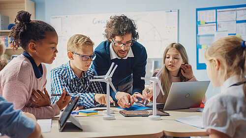 A man explains something to small children and points to a tablet. There are small wind turbine models on the table and laptops and tablets in front of the children.