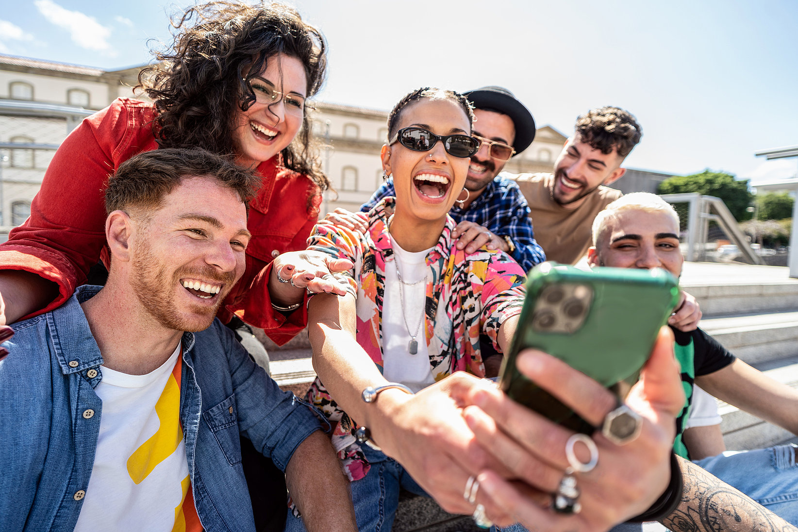  Un grupo de seis amigos están reunidos, sonriendo y riendo mientras se toman un selfie. El ambiente es alegre y el escenario está al aire libre bajo la luz del sol.