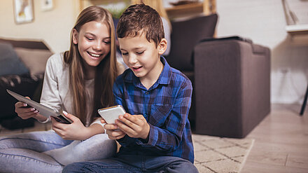 Children's Online Safety A boy with a mobile phone and a girl with a tablet are sitting on the floor in the living room, smiling and showing each other something on it.