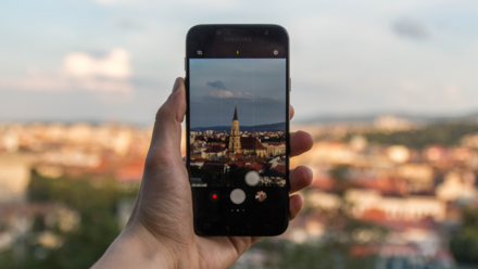 Planear tu viaje haciendo scroll: TravelerZ Una mano sostiene un teléfono capturando la vista de una ciudad al atardecer, combinando tecnología y paisaje en un momento tranquilo y contemplativo.