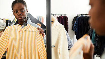 Finanzbildung bei der Next Gen? Teenagerin hält sich im Spiegel eines Einkaufsladens ein Oberteil an.