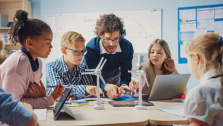 Research News: A new study into the use of technology in secondary schools A man explains something to small children and points to a tablet. There are small wind turbine models on the table and laptops and tablets in front of the children.