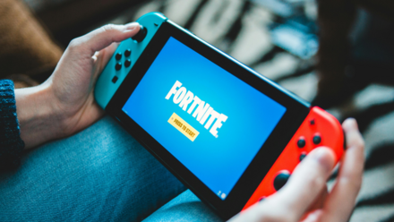 El Gaming como nuevo territorio de Marketing Persona sosteniendo una Nintendo Switch, mostrando la pantalla de título del juego «Fortnite» con la indicación «Pulsar para empezar.» La consola tiene mandos Joy-Con de color azul brillante y rojo.