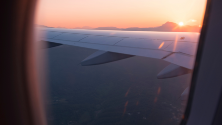 La Generación Z no coquetea con la idea de viajar, ¡la GenZ va directa! Vista de un ala de avión desde la ventana durante el amanecer o atardecer, con un cielo colorido y montañas en el horizonte, evocando tranquilidad y viaje.