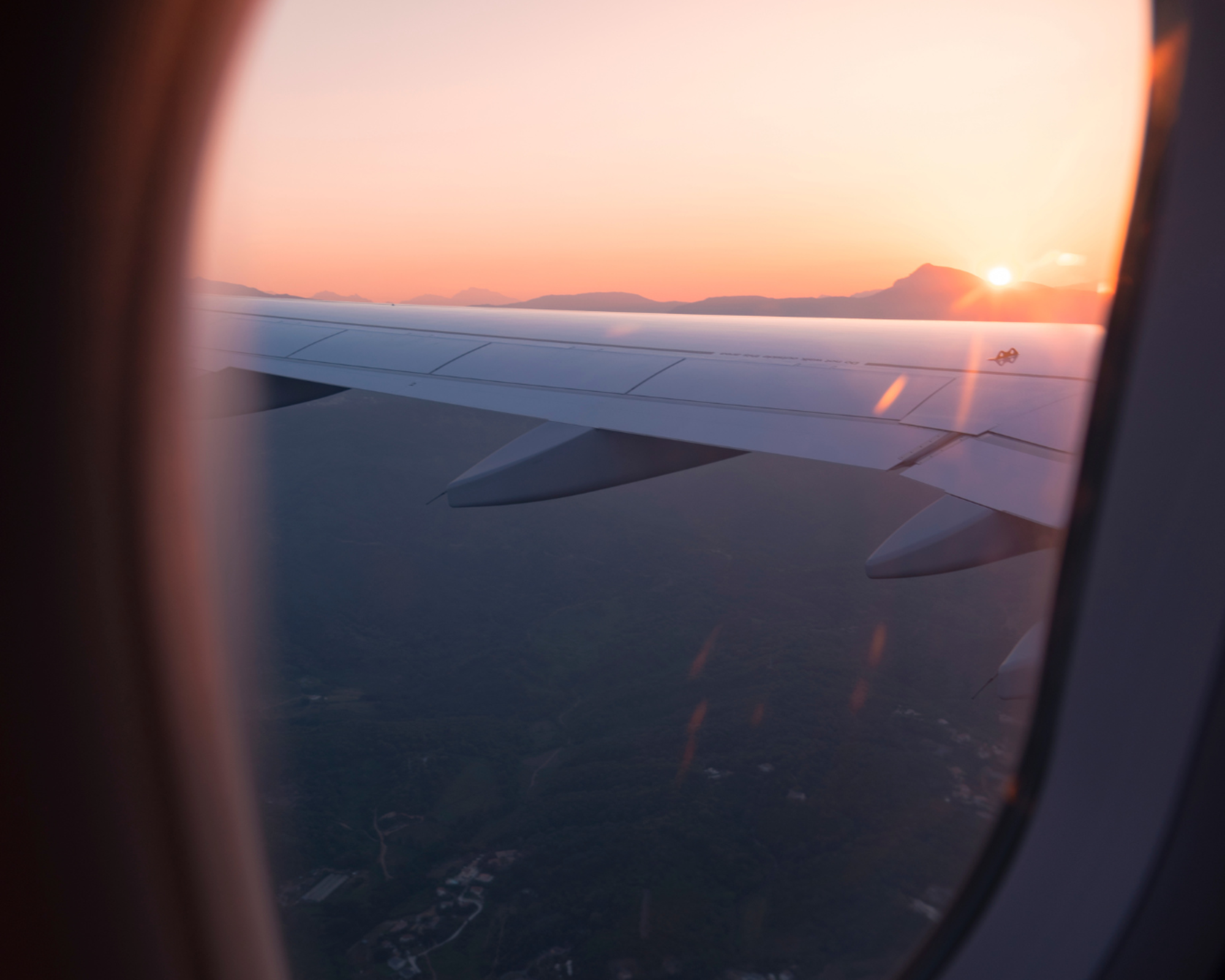 Vista de un ala de avión desde la ventana durante el amanecer o atardecer, con un cielo colorido y montañas en el horizonte, evocando tranquilidad y viaje.