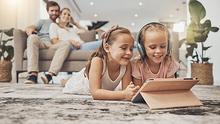 Publicidad kid-oriented: conoce las claves del éxito para 2024 Dos niñas están tumbadas en el suelo, una de ellas con auriculares, mientras ven algo en una tableta. Al fondo, sus padres están sentados en el sofá, sonriendo y observando.