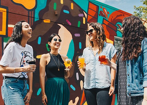 Four women are standing outside with a drink in their hands. The sun is shining and everyone is smiling.