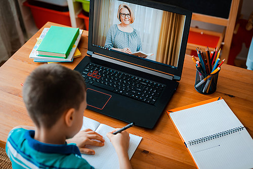 A little boy is doing homework while the lap top is on the table in front of him and a teacher can be seen on the screen.