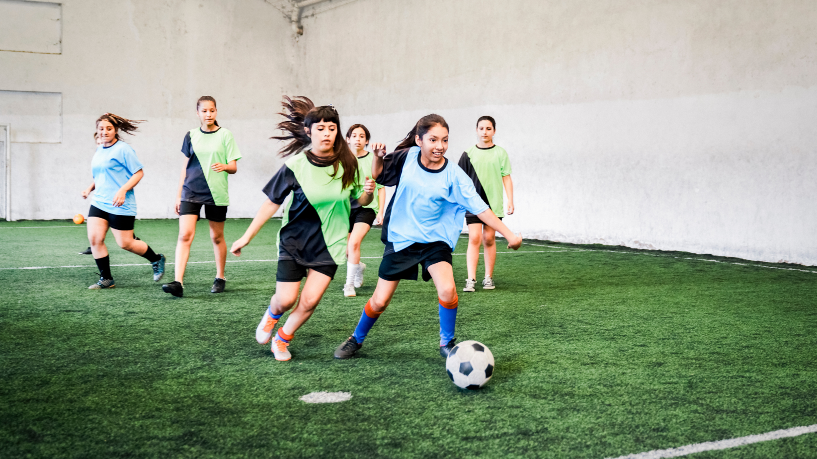 Girls are playing soccer in a hall, with two teams wearing different shirts in blue and green. One girl in the blue jersey dribbles the ball while the others try to catch it.