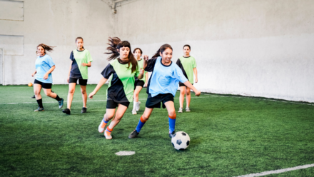 Empowering Tomorrow’s Athletes: The Quiet Revolution in Girls' Sports Participation Girls are playing soccer in a hall, with two teams wearing different shirts in blue and green. One girl in the blue jersey dribbles the ball while the others try to catch it.