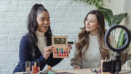 Influencer Marketing: 5 puntos clave para su implementación efectiva Dos mujeres sentadas juntas, una con una paleta de sombras de ojos abierta y la otra mirando. Parecen estar grabando un tutorial de maquillaje, con productos de belleza y un anillo de luz sobre la mesa.