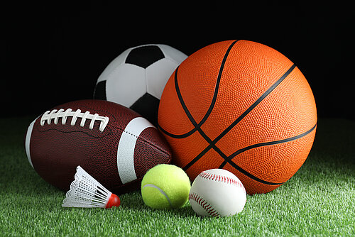 Several sports balls and a shuttlecock lie on green artificial turf against a black background.