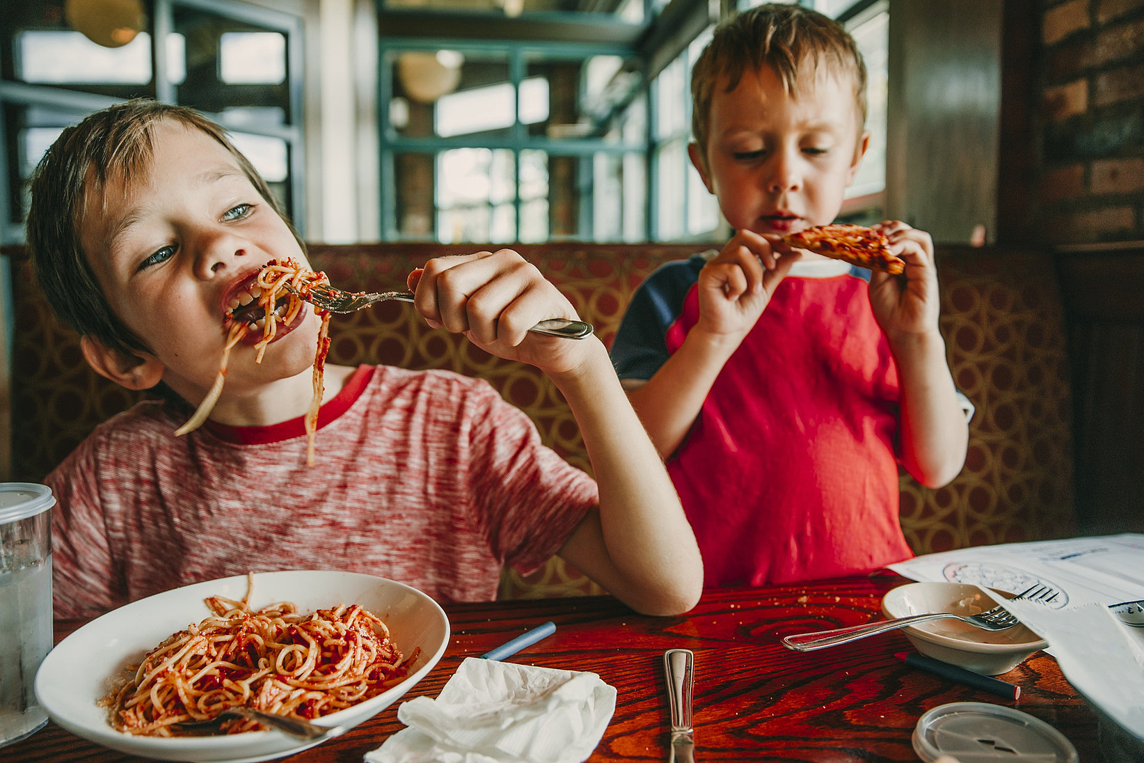 Two little boys are eating spaghetti and pizza at the table.