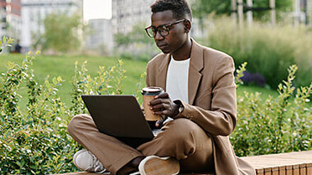 Junger Mann in Businesskleidung sitzt mit Kaffee und Laptop im Freien.