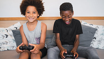 Five Things Games Can Do Beyond Just Entertain Two boys sit smiling on a sofa while they play a game.