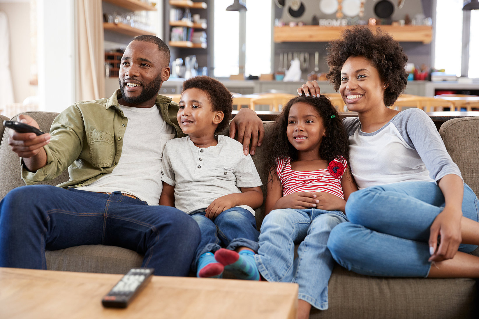A family with two children watches TV on the couch and smiles.