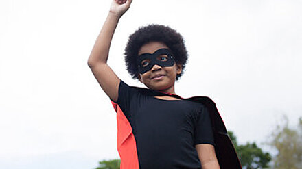 Perspektivwechsel Lächelndes Kind in Superheldenverkleidung streckt den Arm in die Luft.