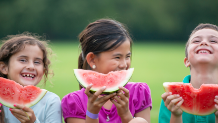 Food&Me: Tendencias en la alimentación de nuestros jóvenes Tres niños sentados al aire libre, sonriendo y disfrutando de rebanadas de sandía en un día soleado, capturando un alegre momento de diversión veraniega.
