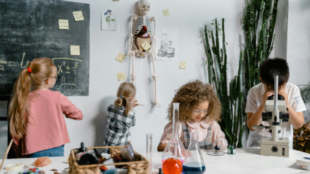 ¿Qué pueden aportar las empresas a la Comunidad Educativa? Los niños participan en una actividad científica en un aula, examinando un modelo de esqueleto, escribiendo en una pizarra y utilizando un microscopio, fomentando la curiosidad y el aprendizaje práctico.