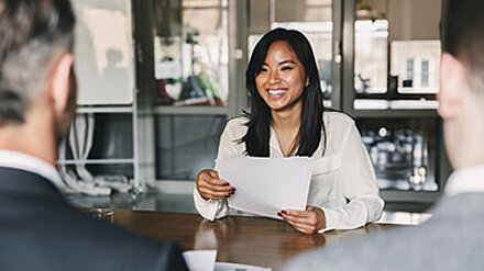Wie überzeugen Young Candidate Journeys wirklich? Junge Frau sitzt lächelnd im Bewerbungsgespräch mit ihren Unterlagen in der Hand.