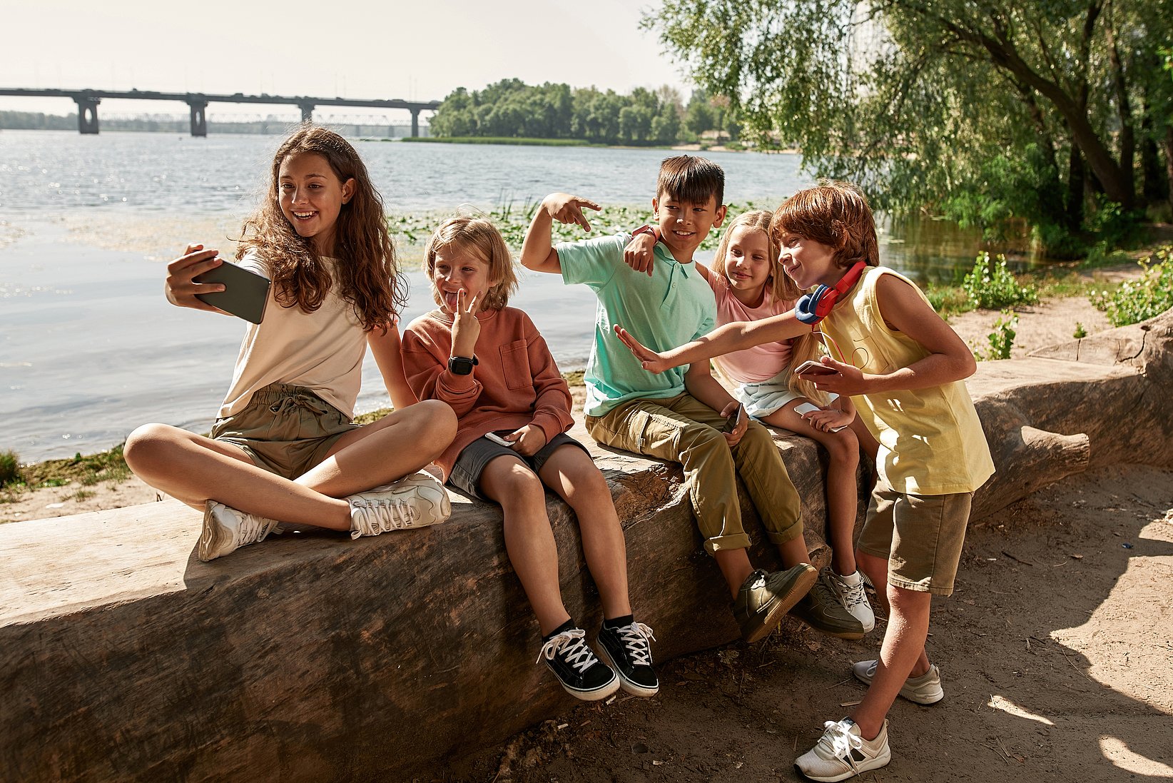 Two girls and three boys are sitting on a wall and making different faces while a girl takes the picture.