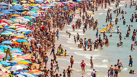 "Overtourism" y "Turismofobia": Los Desafíos Actuales en el Turismo Global Una escena de playa abarrotada con numerosas personas en el agua y coloridas sombrillas cubriendo la arena, representando un ajetreado día de verano junto al mar.