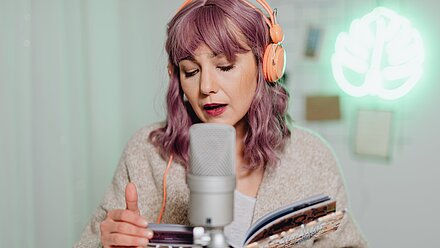 ¿Sigue siendo el podcast una tendencia en marketing? Mujer con auriculares naranjas, cabello morado y un micrófono frente a ella, leyendo en voz alta un libro. El fondo incluye una luz decorativa de neón con forma de hoja.