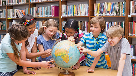 TWEEN COUNCIL: nuestro panel internacional de investigación Un grupo de niños se reúne en torno a un globo terráqueo en una biblioteca, señalando distintos lugares y hablando de geografía. Parecen interesados y curiosos mientras exploran juntos el mapa.