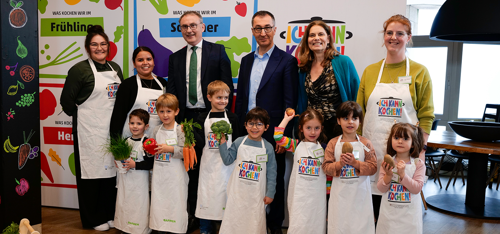 A group of adult women and men in the background and small children (girls and boys) wearing a white apron with the words "I can cook" and smiling at the camera. The children are holding up various vegetables. 