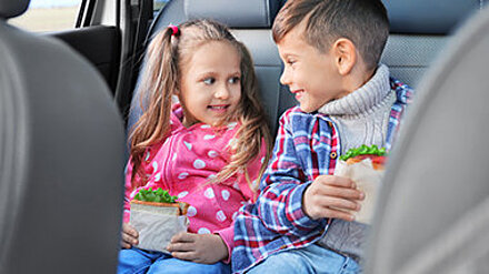 Aufwachsen zwischen Snacks und Mini-Mahlzeiten? Mädchen und Junge sitzen auf der Rückbank eines Autos und genießen lächelnd ihre Pausenbrote.