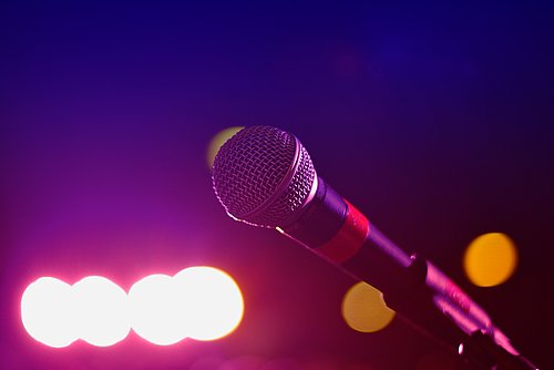 A microphone in the foreground, while it is dark but colourful lights can be seen in the back.