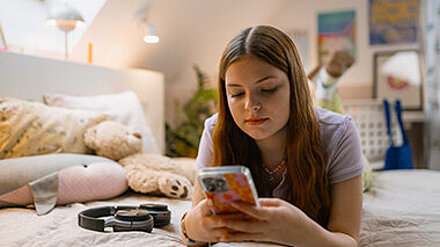 Teenagerin liegt auf dem Bett und sieht in ihr Smartphone.