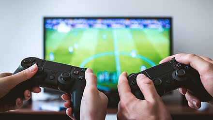 ¿Son los videojuegos un entretenimiento o un escape emocional para la Gen Z? Dos personas juegan un videojuego en una consola con controles negros, mientras una pantalla muestra un partido de fútbol virtual. El enfoque está en las manos y los controles.
