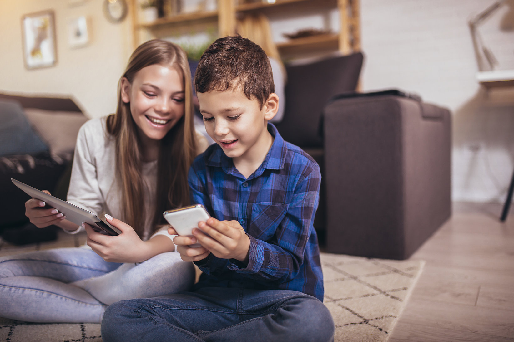 A boy with a mobile phone and a girl with a tablet are sitting on the floor in the living room, smiling and showing each other something on it.