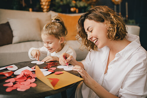 Mother and daughter happily tinker together at a table with colorful paper hearts and decorations.