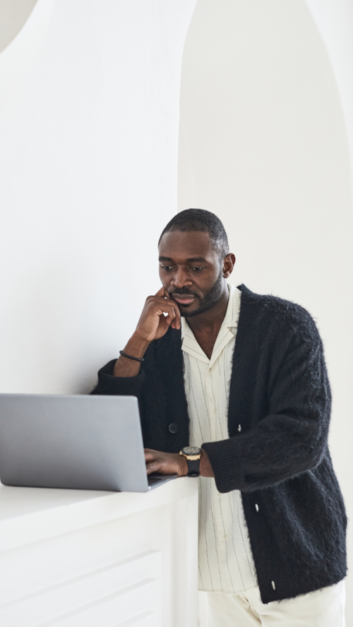 A young man sits at a table in front of his laptop and looks at the screen.
