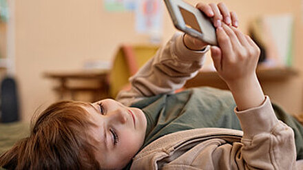 Hey Alexa, wie tickt eigentlich Gen A? Kind liegt auf dem Rücken in seinem Kinderzimmer und schaut in ein Smartphone.