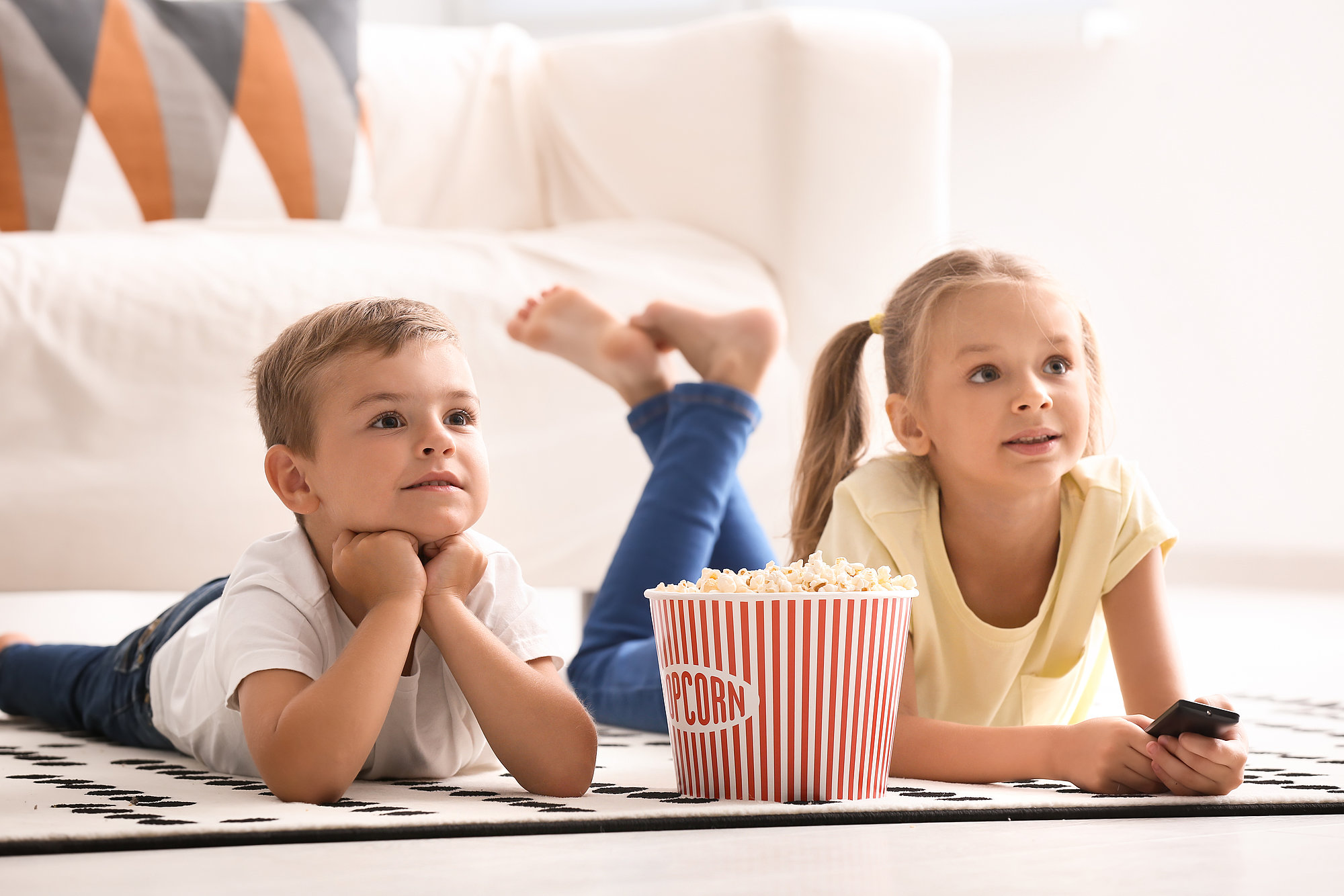  Two young children lying on the floor, watching TV with a large bucket of popcorn in front of them.