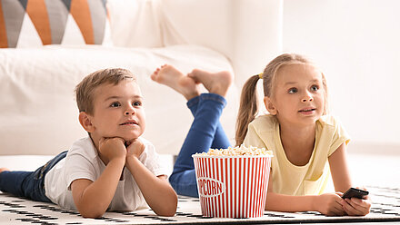 Kids TV Shows We Are Excited for in 2024 Two young children lying on the floor, watching TV with a large bucket of popcorn in front of them.