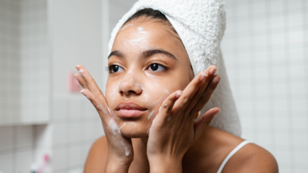 Tendencias de Belleza 2024: Guía para Conectar con la Gen Z Una joven con una toalla en la cabeza se aplica un limpiador facial mientras se mira en el espejo, concentrada en su rutina de cuidado de la piel.