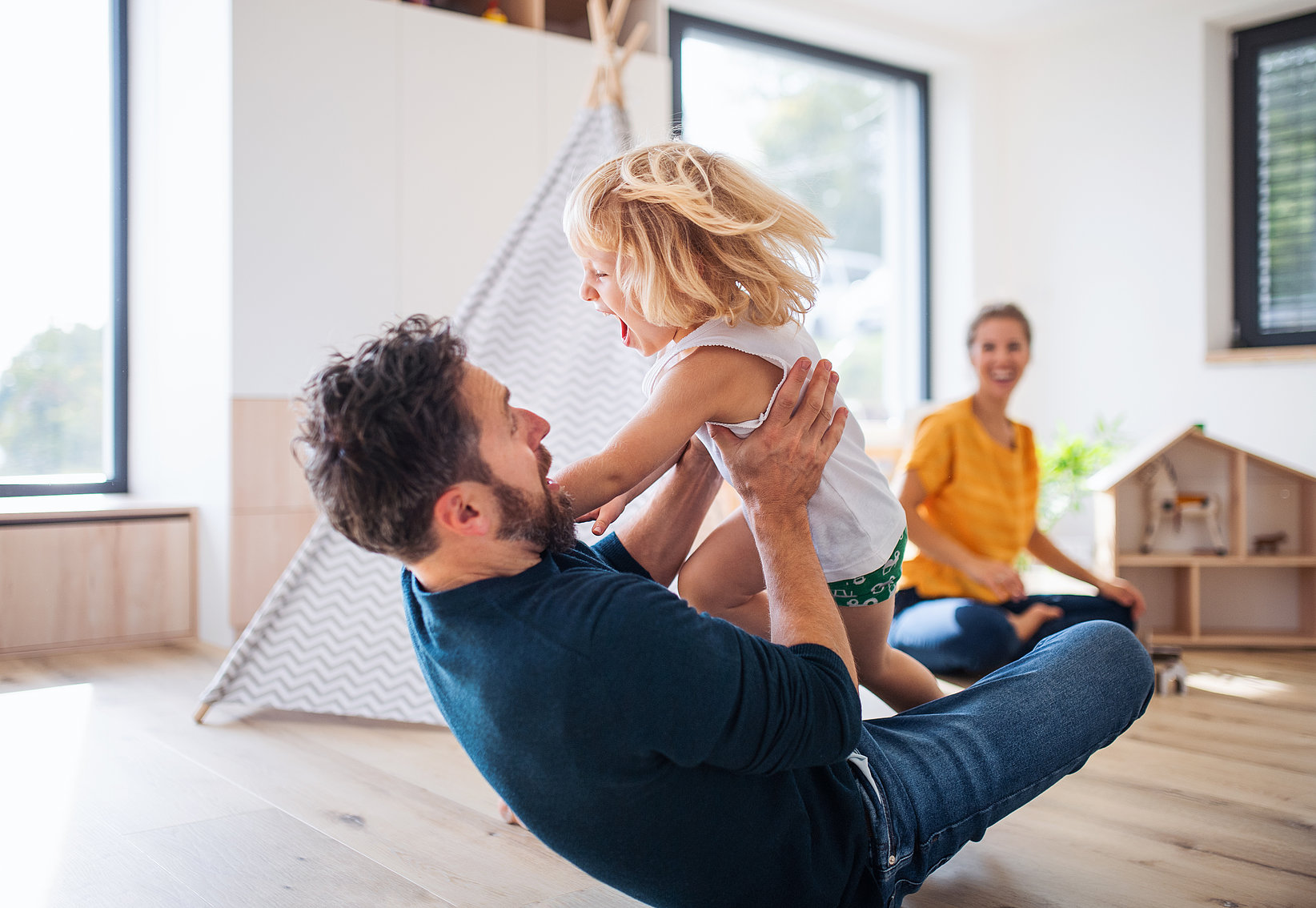 Un padre juega con su hijo pequeño, levantándolo en el aire, mientras el niño ríe alegremente. Al fondo, una madre sonriente observa, creando un cálido momento familiar en un hogar luminoso y juguetón.