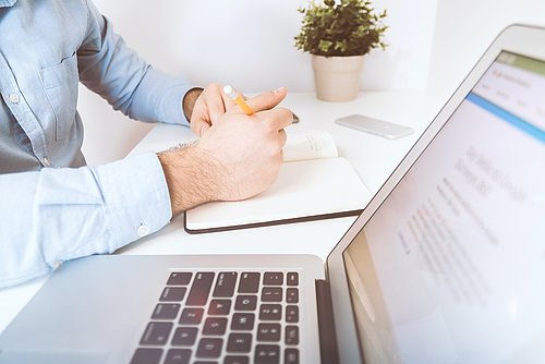 Person writes in a notebook while a laptop is open on the desk.