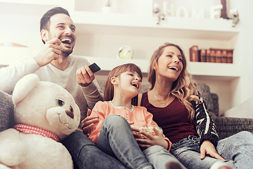 Parents and their daughter are sitting on the couch, watching TV and laughing.