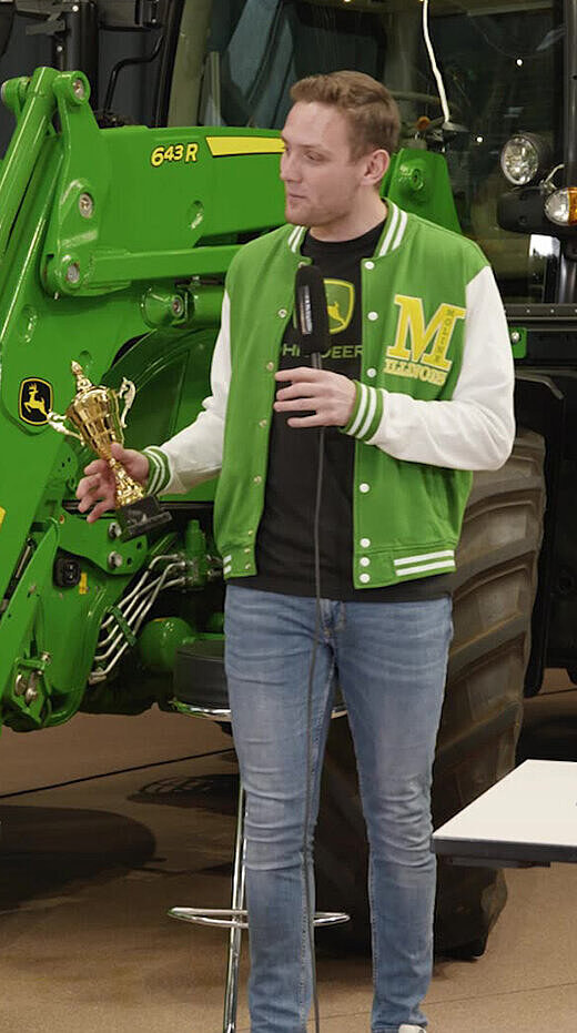 Mann in grüner Jacke steht vor einem grünen Traktor und erklärt etwas.