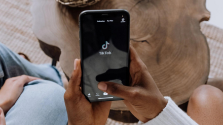 TikTok revoluciona la forma en que los jóvenes consumen contenido de larga duración Una persona sostiene un smartphone y muestra la aplicación TikTok en la pantalla. El teléfono está enfocado, mientras que el fondo incluye una superficie de madera y la mano de la persona.