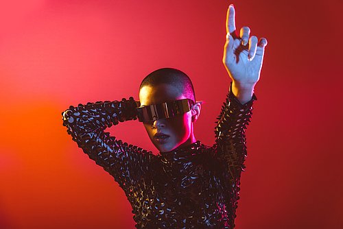 Woman wears a shiny costume, sunglasses and stretches out her arm as if doing a dance step. The background is red and orange.