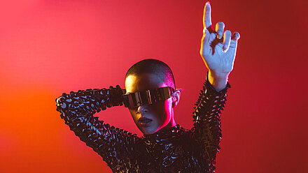 The Rise of Virtual Influencers Woman wears a shiny costume, sunglasses and stretches out her arm as if doing a dance step. The background is red and orange.