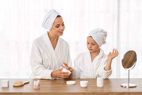 Mother and daughter in white bathrobes stand together and apply skincare products. The scene conveys a feeling of relaxation and a shared self-care routine.