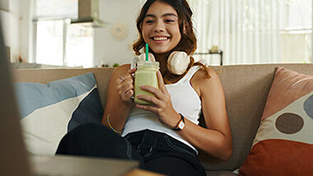 Teenagerin sitzt lächelnd auf dem Sofa und trinkt einen Smoothie.