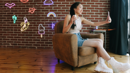 Hooks: La Clave para capturar la atención con tus publicaciones Una joven está sentada en una acogedora silla, con una copa en la mano y tomándose un selfie contra una pared de ladrillo decorada con coloridos carteles de neón y las palabras «Rock Star». La escena desprende un ambiente divertido y vibrante.